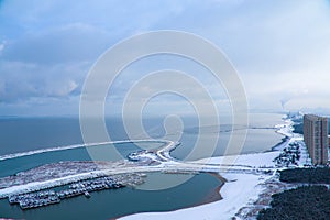 A distant view of the harbor. A bustling city sits along the coastline. The port is filled with ships and covered in heavy snow.