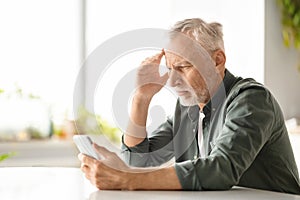 Distressed elderly man suffering headache while looking at smartphone at home