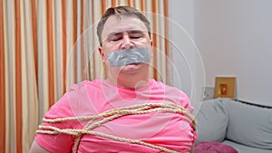 distressed Man in pink shirt grimacing tied with ropes and mouth sealed with duct tape, hinting at an abduction scenario