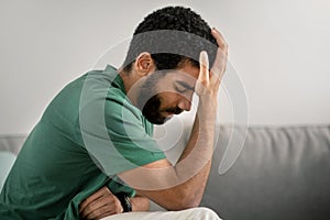 Distressed young man with a beard, dressed in a green t-shirt, sitting on a grey sofa