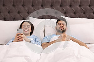 Distrustful young man peering into girlfriend`s smartphone in bed