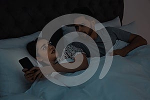 Distrustful young man peering into girlfriend`s smartphone in bed