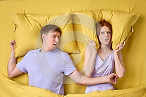 Disturbed Young Woman Covering Ears With Pillows While Man Snoring On Bed