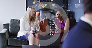 Diverse businesswomen sitting using laptop drinking coffee in office lounge