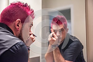 Diverse caucasian man with spiked pink hair applying eyeliner in the mirror