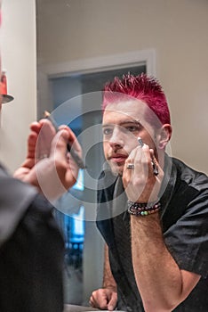 Diverse caucasian man with spiked pink hair applying eyeliner in the mirror