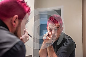 Diverse caucasian man with spiked pink hair applying eyeliner in the mirror