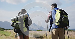Diverse couple wearing backpacks using nordic walking poles hiking in countryside