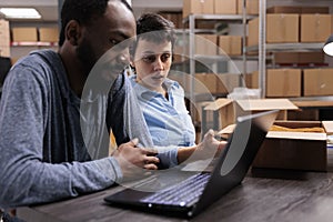 Diverse employees sitting at table in warehouse checking transportation logistics on laptop computer