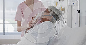 Diverse female doctor giving oxygen mask to senior female patient in hospital bed, slow motion