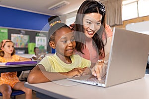 Diverse female teacher and schoolgirl using laptop in elementary school class