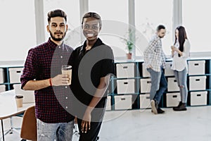 Diverse friends two coworkers standing at office distracted from work take break drink coffee having informal talk. Boss employee