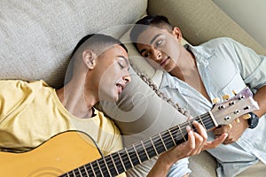 Diverse gay male couple lying on sofa playing guitar