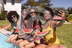 Diverse group of female friends taking selfie with watermelon sitting at the poolside