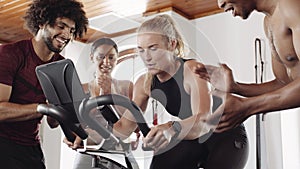 Diverse group of fit young adults cheering around Caucasian female friend while she cycles on stationary exercise bike