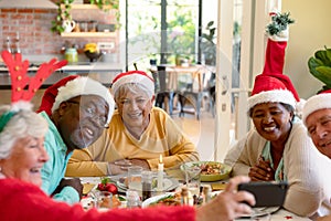 Diverse group of happy senior friends in holiday hats celebrating christmas together, taking selfie