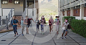 Diverse group of schoolchildren wearing backpacks and face masks running at school yard