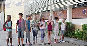 Diverse group of schoolchildren wearing backpacks smiling and standing in a row at school yard
