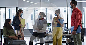 Diverse group of work colleagues wearing face mask holding laptop, tablet and documents talking