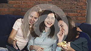 Diverse happy friends sharing pizza together. Women sitting on sofa, hugging and laughing, looking at laptop screen