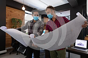 Diverse male colleagues wearing masks in disucssion holding architectural plans in office