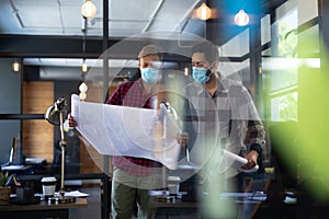 Diverse male colleagues wearing masks in disucssion holding architectural plans in office