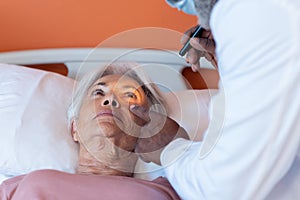 Diverse male doctor examining eye of senior female patient lying on bed with penlight, copy space