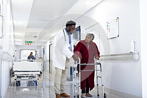 Diverse male doctor helping senior female patient use walking frame in hospital corridor, copy space