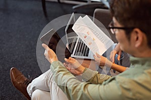 Diverse male and female colleague talking in office lounge holding paperwork using tablet and laptop