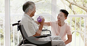 Diverse male physiotherapist advising and senior man in wheelchair using dumbbells, in slow motion