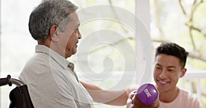 Diverse male physiotherapist advising and senior man in wheelchair using dumbbells, in slow motion