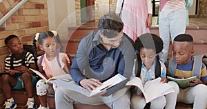 Diverse male teacher and children sitting on stairs reading books in elementary school, slow motion