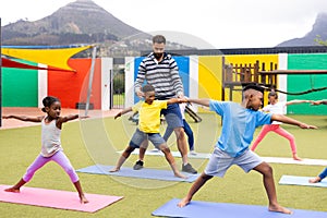 Diverse male teacher and elementary schoolchildren learning yoga in schoolyard