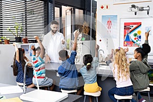 Diverse male teacher and elementary schoolchildren raising hands in biology class