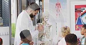 Diverse male teacher and happy schoolchildren studying skeleton in biology class