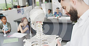 Diverse male teacher and happy schoolchildren studying skeleton in biology class