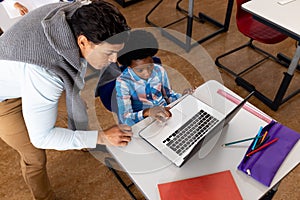 Diverse male teacher using laptop teaching boy in class at elementary school