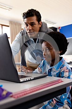 Diverse male teacher using laptop teaching boy in class at elementary school
