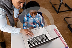 Diverse male teacher using laptop teaching boy at desk in elementary school class