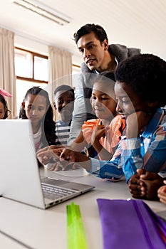 Diverse male teacher using laptop teaching children in class at elementary school