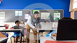 Diverse male teacher using laptop teaching girl in class at elementary school