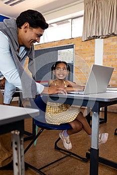 Diverse male teacher using laptop teaching girl in class at elementary school