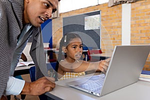 Diverse male teacher using laptop teaching girl in class at elementary school