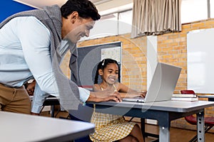 Diverse male teacher using laptop teaching girl at desk in elementary school class