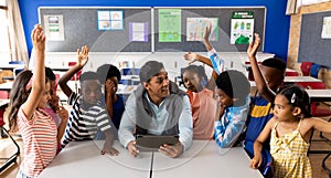 Diverse male teacher using tablet with children raising hands in class at elementary school