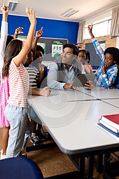 Diverse male teacher using tablet with children raising hands in class at elementary school