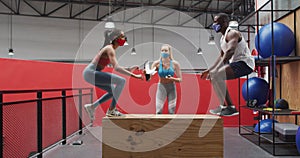 Diverse man and woman jumping on box wearing face masks at gym, caucasian woman timing