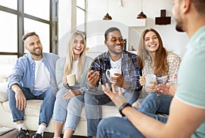 Diverse millennial friends talking and laughing together, drinking tea or coffee at home