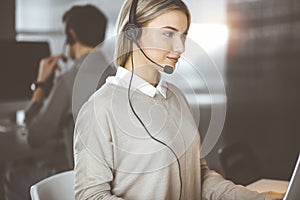 Diverse people group in sunny call center. Blonde business woman talking by headset while sitting in modern office