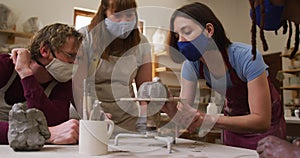 Diverse potters wearing face masks and aprons working on pot on potters wheel at pottery studio